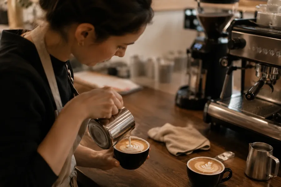 Kursteilnehmerin gießt Milch aus der Kanne in eine Tasse, Latte-Art-Muster entsteht